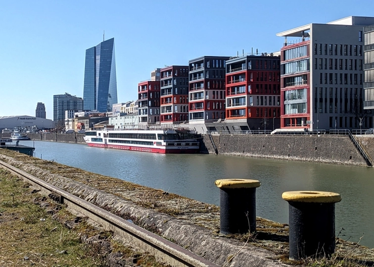 Blick über den Frankfurter Osthafen auf moderne Wohngebäude, ein Flusskreuzfahrtschiff und den markanten Turm der Europäischen Zentralbank im Hintergrund.