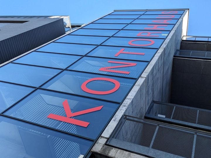 Untersicht einer modernen Glasfassade mit dem vertikalen, roten Schriftzug „KONTORHAUS“ vor blauem Himmel.
