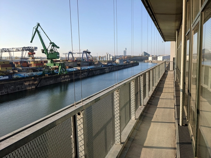 Blick von einem schmalen Industriebalkon auf ein Hafenbecken mit einem großen grünen Verladekran und gestapelten Containern.