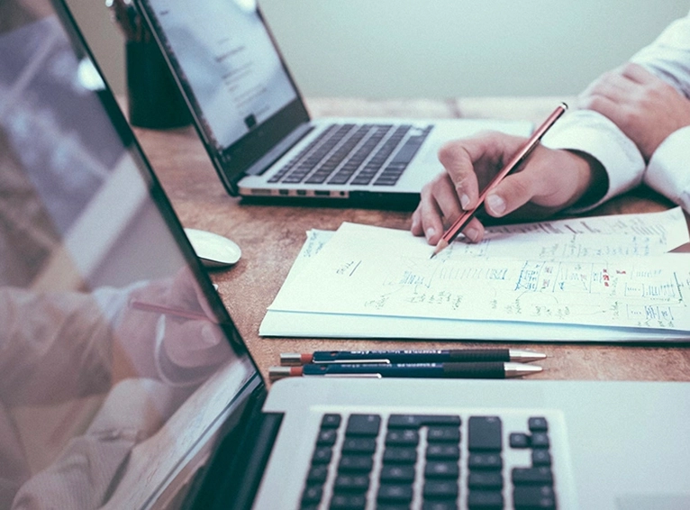 Zwei Personen arbeiten an Laptops. Eine Person schreibt Notizen auf Papier. Holztisch mit Technik und Dokumenten.