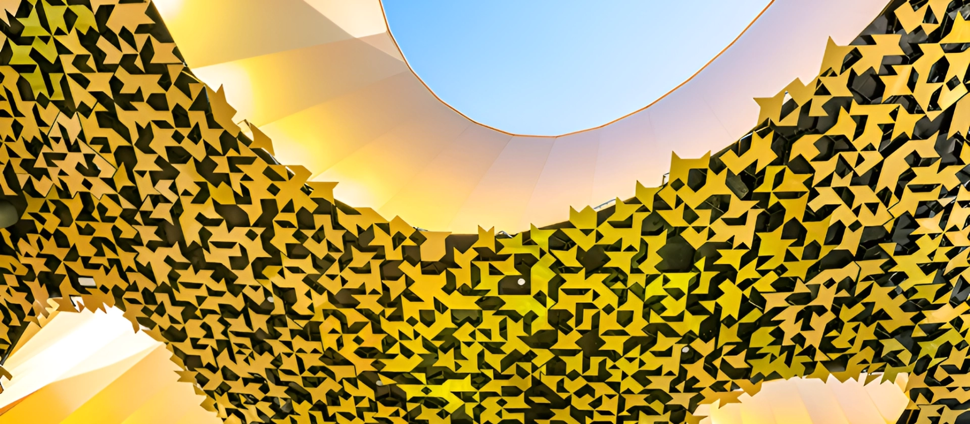 Blick nach oben auf die einzigartige, goldgelbe Decke des House of Music in Budapest, deren geometrische Öffnungen ein rundes Oberlicht mit blauem Himmel freigeben.