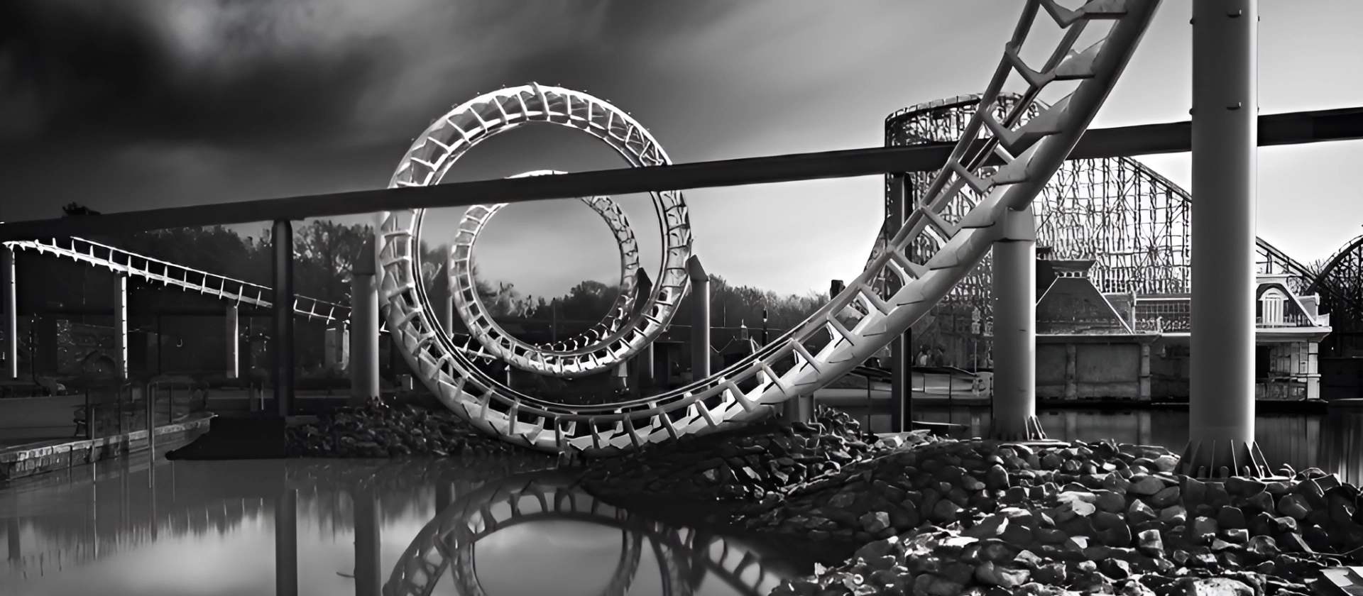 chwarzweißaufnahme der Achterbahn Big Loop im Heide Park Resort in Soltau, mit markanter Looping-Struktur, die sich im ruhigen Wasser davor spiegelt.