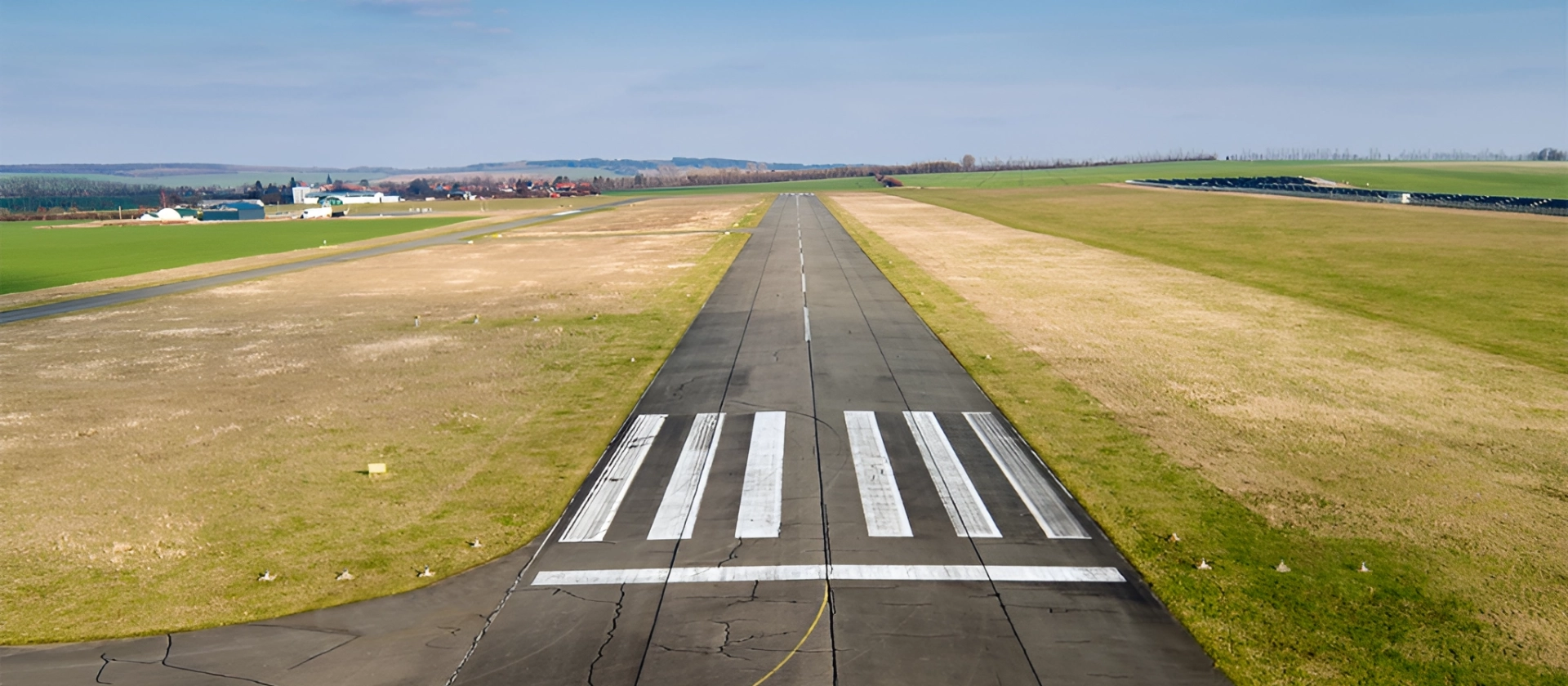 Gerade Start- und Landebahn eines kleinen Flugplatzes, mittig ausgerichtet, umgeben von flachen Wiesen und Feldern unter blauem Himmel mit leichter Bewölkung.