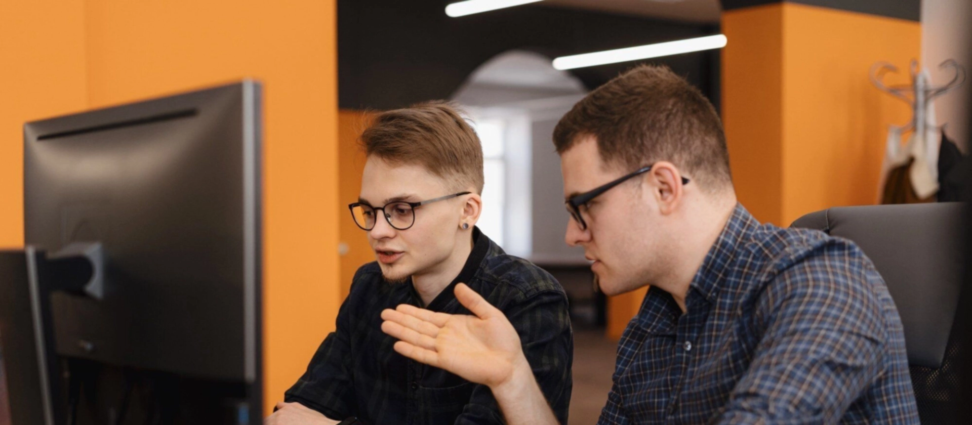 Zwei Kollegen sitzen im Büro vor einem Monitor und besprechen gemeinsam etwas am Computer; einer zeigt gestikulierend auf den Bildschirm