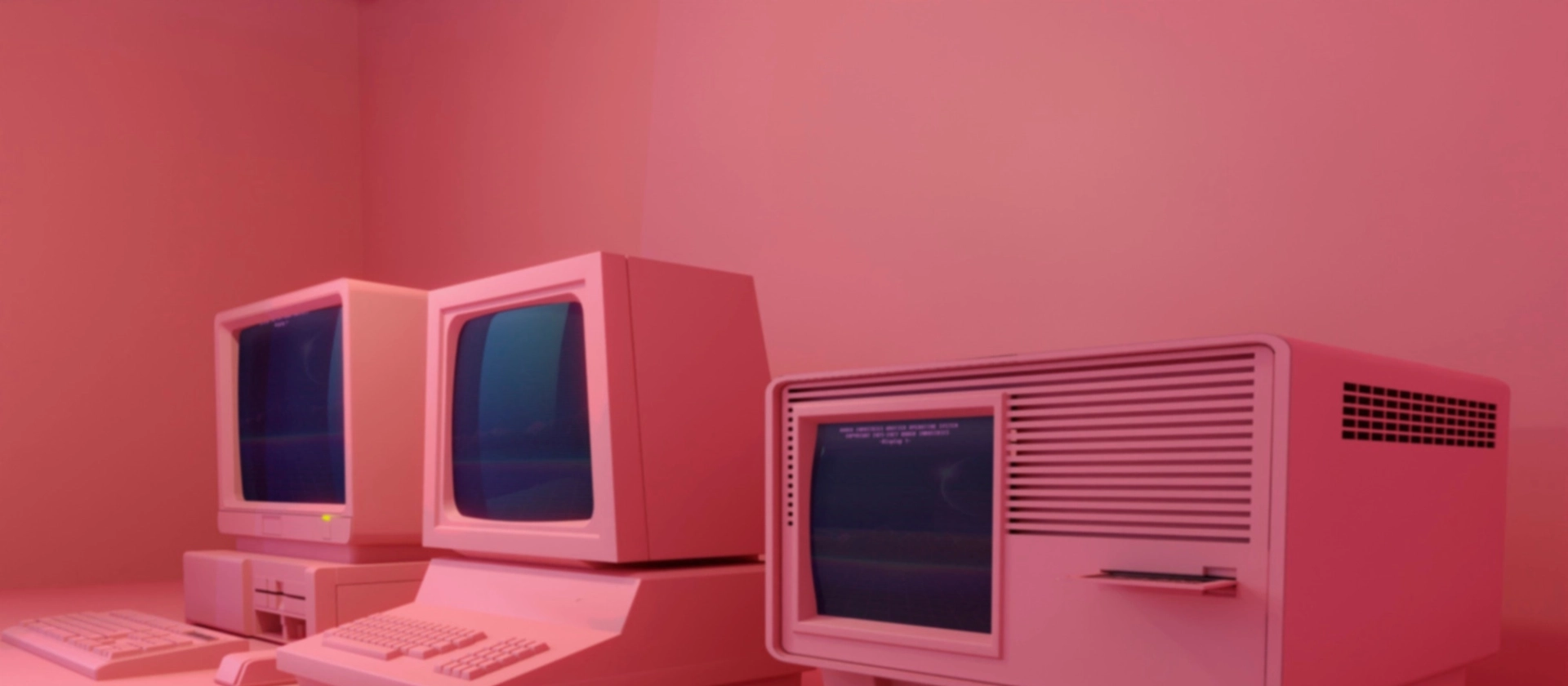 Drei retroartige Computer mit Röhrenmonitoren und Tastaturen stehen nebeneinander auf einem Tisch in einem rosa beleuchteten Raum mit minimalistischem Hintergrund.“