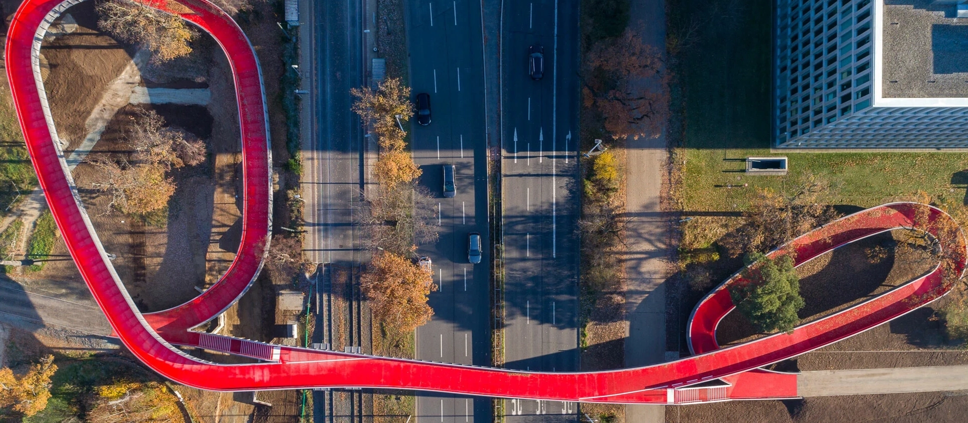 Luftaufnahme einer geschwungenen roten Fuß- und Radwegbrücke über die mehrspurige Rheinstraße in Darmstadt, umgeben von Bäumen, Wegen und Gebäuden.