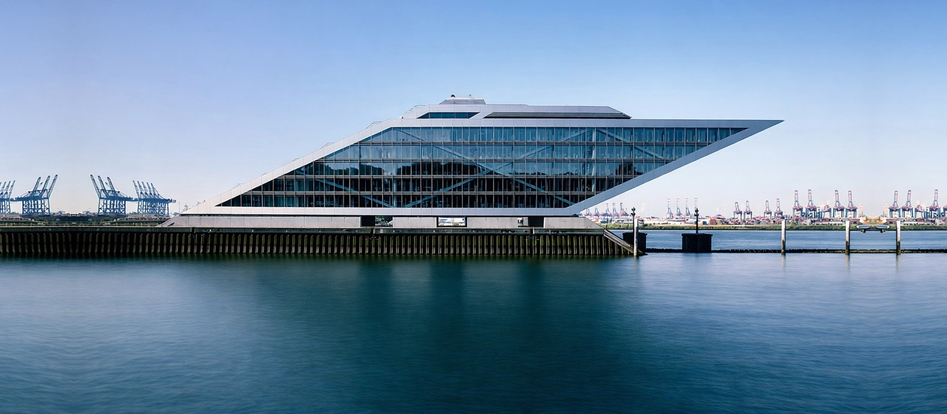 Moderne, keilförmige Glasarchitektur an einem Hafenbecken, spiegelnde Fassade über ruhigem Wasser, im Hintergrund Kräne und klarer blauer Himmel.