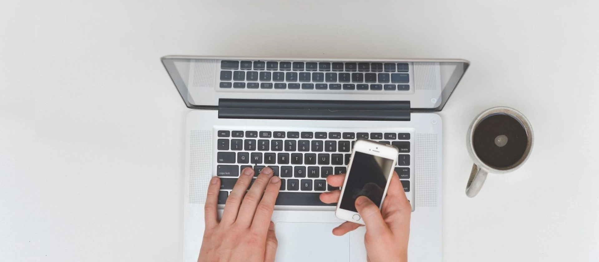 Blick von oben auf eine Person, die an einem Laptop arbeitet und gleichzeitig ein Smartphone hält, neben einer Kaffeetasse auf einer hellen, aufgeräumten Arbeitsfläche