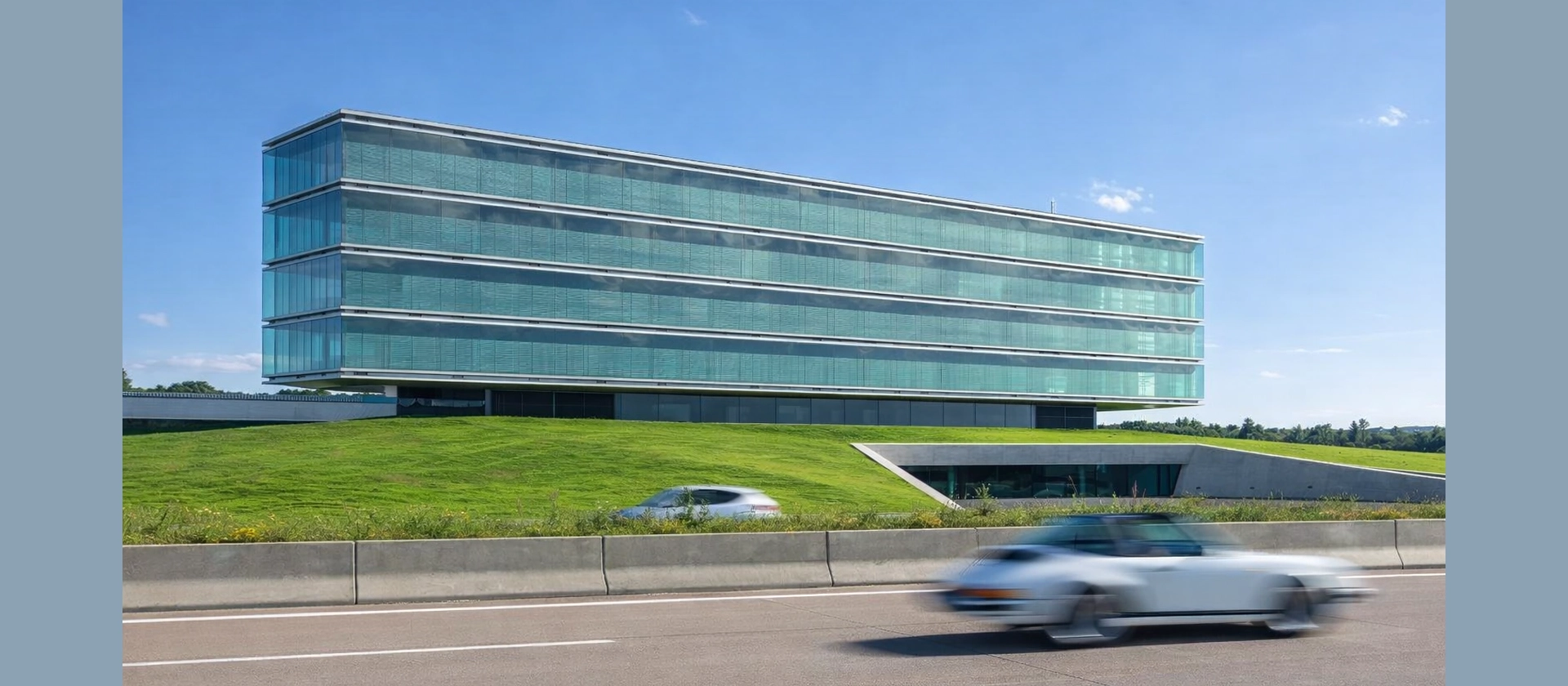 Moderne Firmenzentrale von Robatherm in Jettingen-Scheppach mit langgestreckter Glasfassade auf einer begrünten Anhöhe, davor eine Straße mit vorbeifahrenden Autos unter blauem Himmel.“