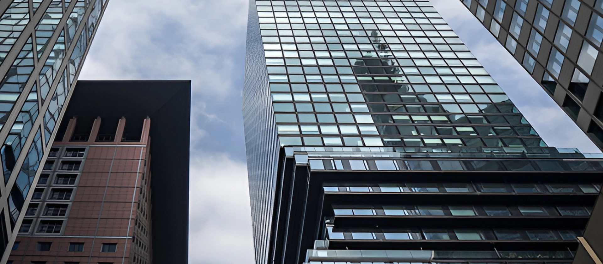 Blick von unten zwischen modernen Hochhäusern mit Glasfassaden und einem Backsteingebäude, die sich gegen einen bewölkten Himmel erheben