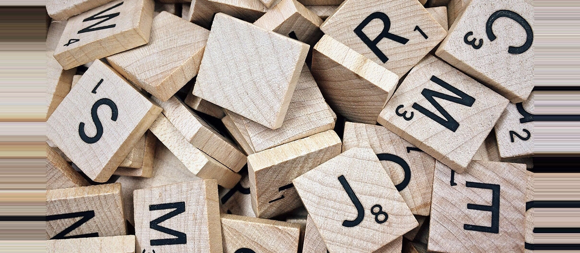 Dicht geschichtete Holzplättchen mit schwarzen Buchstaben und Zahlen liegen ungeordnet beisammen und zeigen eine warme Holzstruktur mit spielerischem Typografiecharakter