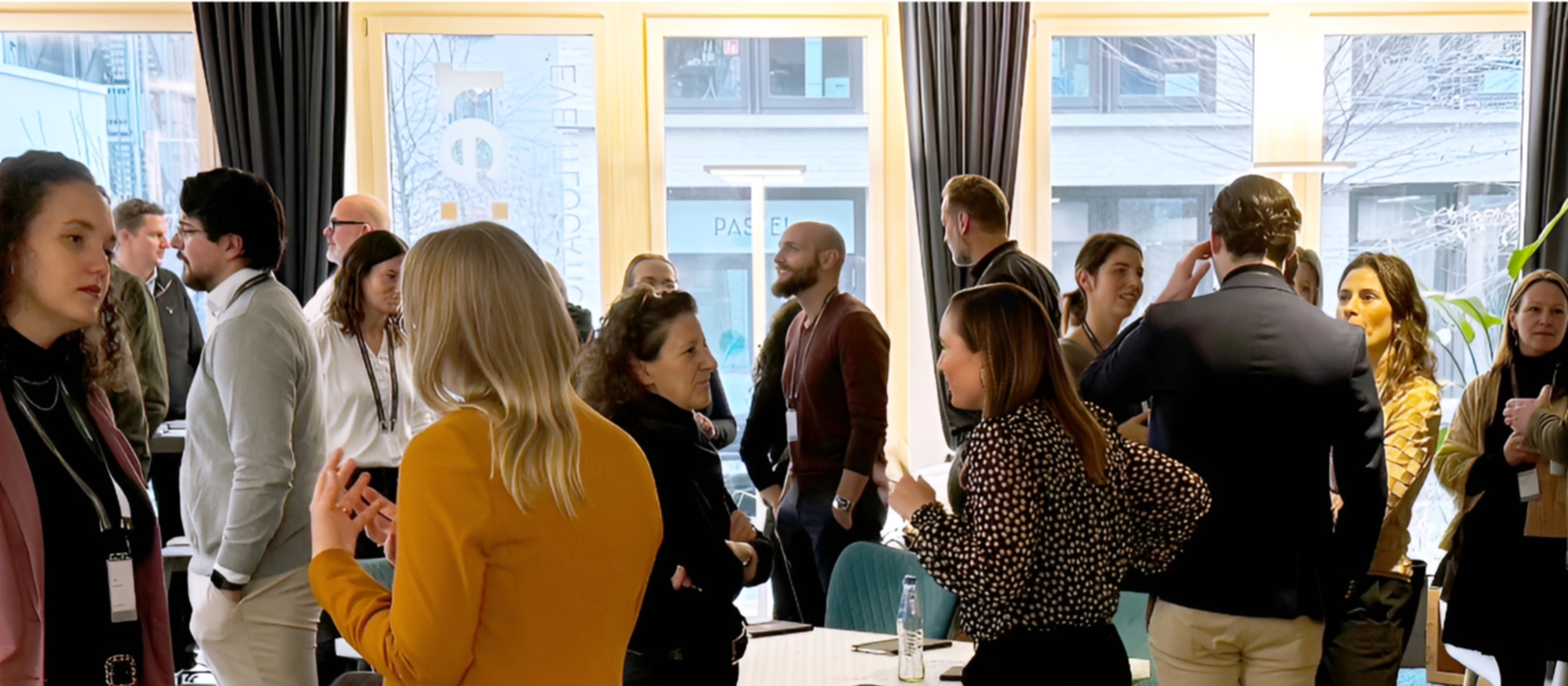 Menschen stehen in einem hellen Raum an Tischen zusammen, unterhalten sich angeregt und netzwerken bei einer Veranstaltung mit Blick durch große Fenster.