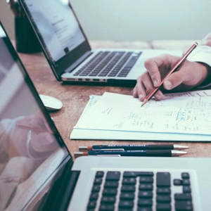 Zwei Personen arbeiten an Laptops. Eine Person schreibt Notizen auf Papier. Holztisch mit Technik und Dokumenten.