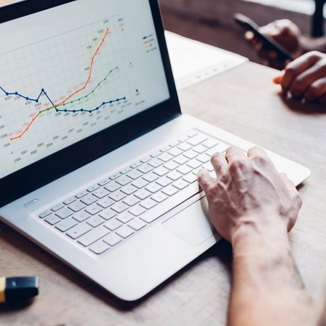 Nahaufnahme einer Person, die auf einem Laptop mit hellem Gehäuse und Tastatur tippt. Auf dem Bildschirm wird ein farbiges Liniendiagramm mit Finanzdaten angezeigt.