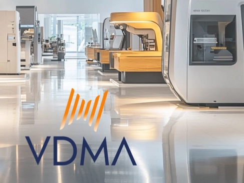 Das Logo des VDMA (Verband Deutscher Maschinen- und Anlagenbau) in Blau und Orange, über einer Aufnahme einer hellen Ausstellungshalle mit modernen Industriemaschinen.