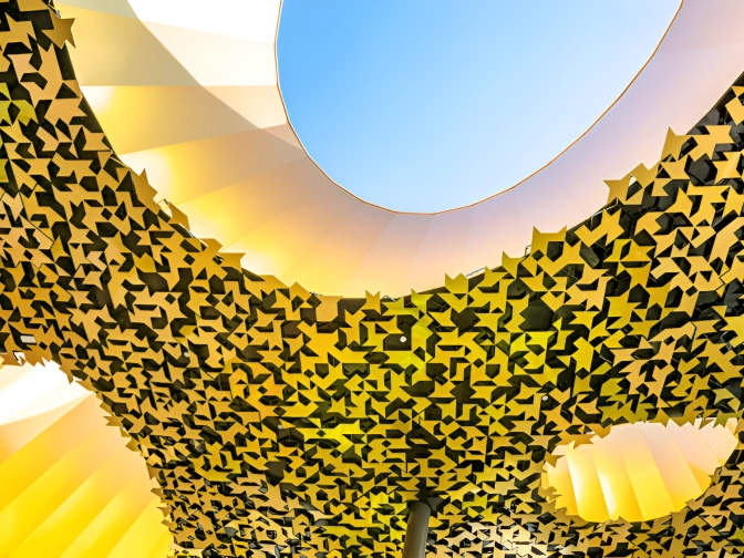 Blick nach oben auf die einzigartige, goldgelbe Decke des House of Music in Budapest, deren geometrische Öffnungen ein rundes Oberlicht mit blauem Himmel freigeben.