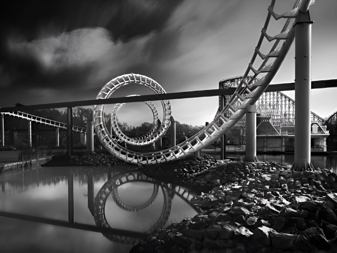 chwarzweißaufnahme der Achterbahn Big Loop im Heide Park Resort in Soltau, mit markanter Looping-Struktur, die sich im ruhigen Wasser davor spiegelt.