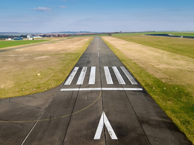 Gerade Start- und Landebahn eines kleinen Flugplatzes, mittig ausgerichtet, umgeben von flachen Wiesen und Feldern unter blauem Himmel mit leichter Bewölkung.
