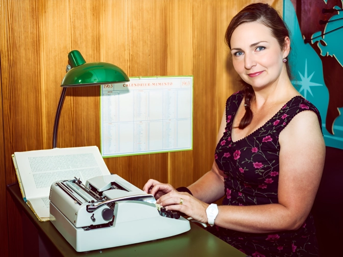 rau sitzt an einer Schreibmaschine an einem Schreibtisch, tippt Text und blickt lächelnd zur Kamera, beleuchtet von einer grünen Schreibtischlampe vor Holzvertäfelung.