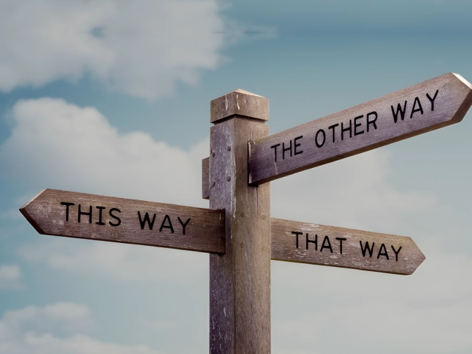 Hölzerner Wegweiser mit drei Pfeilen, beschriftet mit ‚This Way‘, ‚That Way‘ und ‚The Other Way‘, vor blauem Himmel mit vereinzelten Wolken