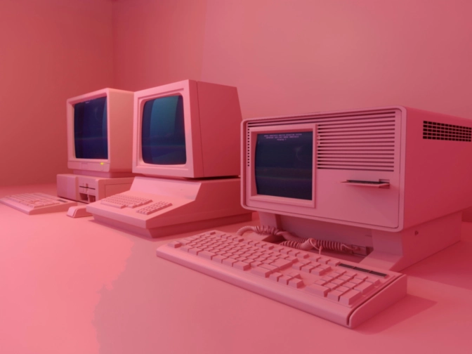 Drei retroartige Computer mit Röhrenmonitoren und Tastaturen stehen nebeneinander auf einem Tisch in einem rosa beleuchteten Raum mit minimalistischem Hintergrund.“