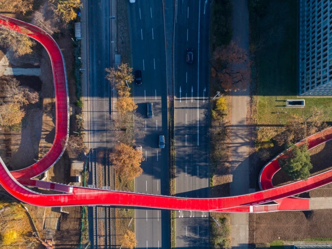 Luftaufnahme einer geschwungenen roten Fuß- und Radwegbrücke über die mehrspurige Rheinstraße in Darmstadt, umgeben von Bäumen, Wegen und Gebäuden.