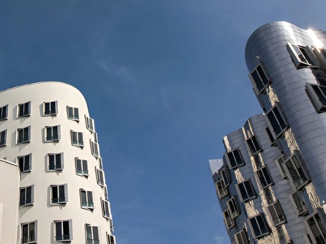 Moderne, geschwungene Bürogebäude mit weißen und metallisch glänzenden Fassaden ragen vor einem klaren, tiefblauen Himmel auf und wirken futuristisch und lichtdurchflutet.“