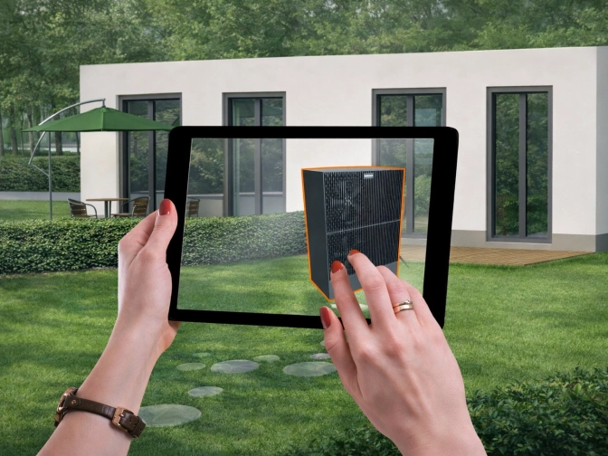 Hände halten ein Tablet im grünen Garten vor einem modernen Wohnhaus, auf dessen Bildschirm ein virtuell platziertes schwarzes Technikgerät zu sehen ist
