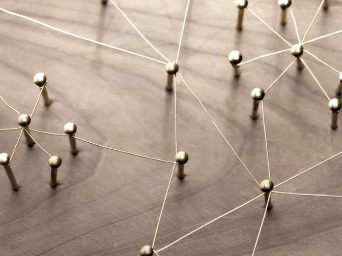 Metallene Stifte mit kugelförmigen Köpfen sind durch feine Drähte zu einem Netzwerk verbunden und liegen auf einer warmen Holzoberfläche, was Vernetzung und Beziehung symbolisiert.