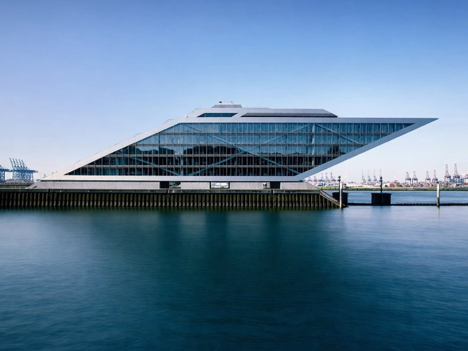 Moderne, keilförmige Glasarchitektur an einem Hafenbecken, spiegelnde Fassade über ruhigem Wasser, im Hintergrund Kräne und klarer blauer Himmel.