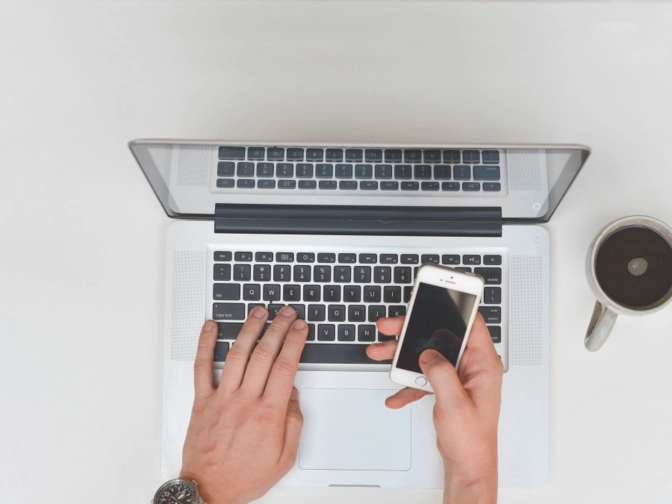 Blick von oben auf eine Person, die an einem Laptop arbeitet und gleichzeitig ein Smartphone hält, neben einer Kaffeetasse auf einer hellen, aufgeräumten Arbeitsfläche
