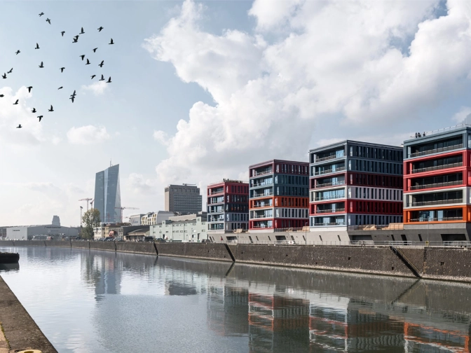 Hafenkana in Frankfurt am Main mit ruhiger Wasseroberfläche, farbigen Bürogebäuden am Ufer, Krananlage links und Vögeln am Himmel unter großen, hellen Wolken