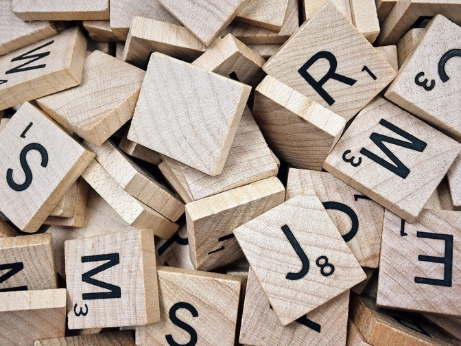 Dicht geschichtete Holzplättchen mit schwarzen Buchstaben und Zahlen liegen ungeordnet beisammen und zeigen eine warme Holzstruktur mit spielerischem Typografiecharakter