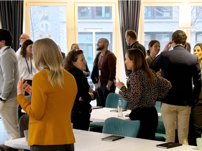 Menschen stehen in einem hellen Raum an Tischen zusammen, unterhalten sich angeregt und netzwerken bei einer Veranstaltung mit Blick durch große Fenster.