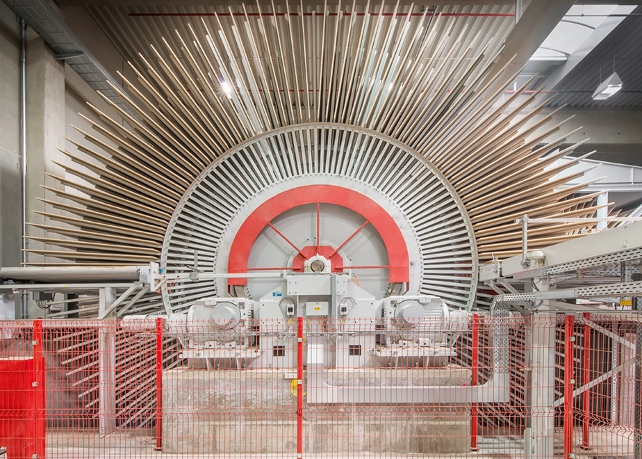 Industrielle Aufnahme einer großen, radialen Maschine in einer Fabrikhalle, deren oberer Halbkreis mit vielen Holzlatten besetzt ist.