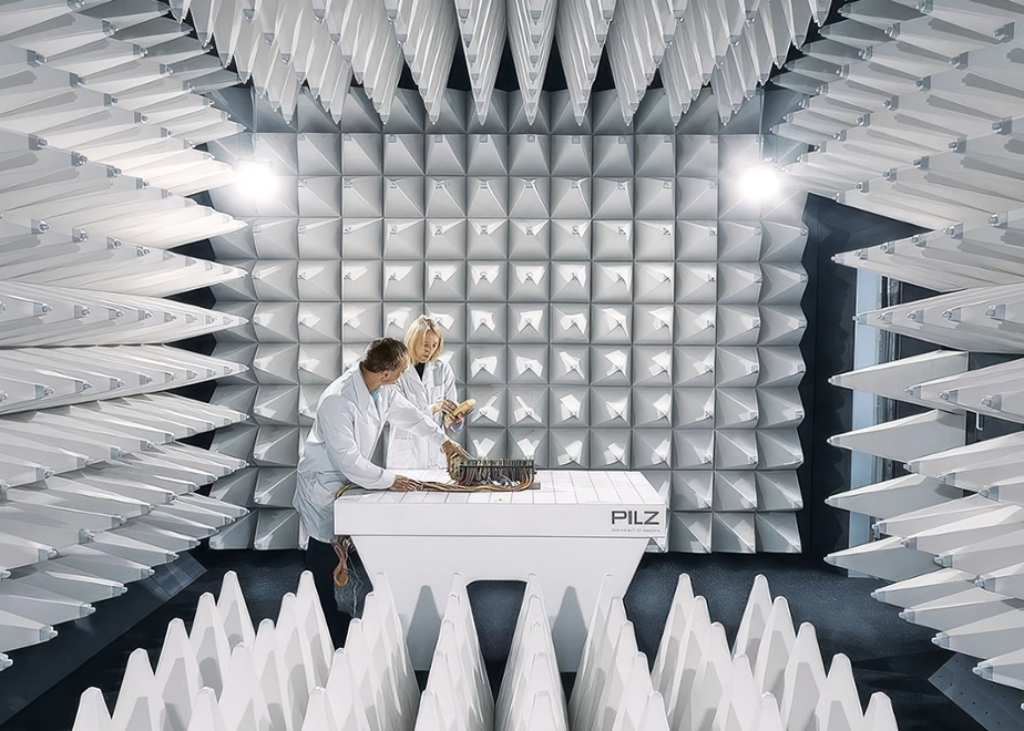 Zwei Techniker in Laborkitteln arbeiten an einem Tisch in einem weißen, pyramidenförmigen EMV-Prüfraum.