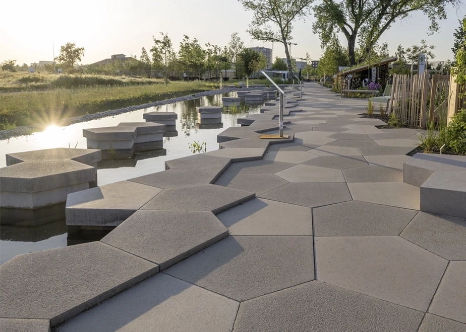 Query successful  Gemini said  Ein moderner Gehweg aus großformatigen, hexagonalen Betonplatten verläuft entlang eines Wasserbeckens mit passenden Trittsteinen in einer sonnigen Parkanlage.
