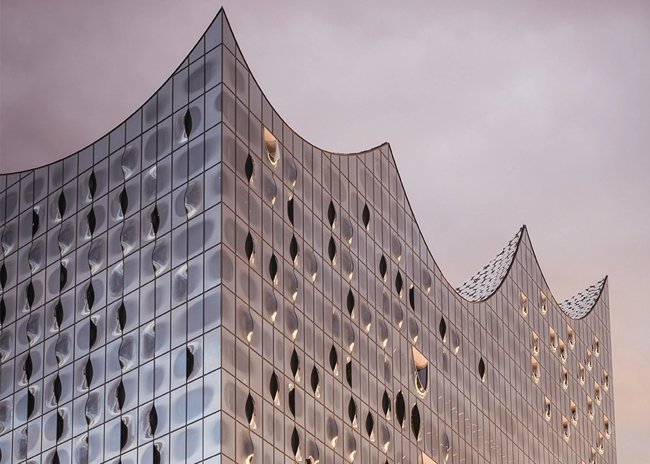 Modern glass building with a distinctive wave-shaped roof located beside a river at sunset. The illuminated interior contrasts with the reflective glass façade, showcasing contemporary architecture.